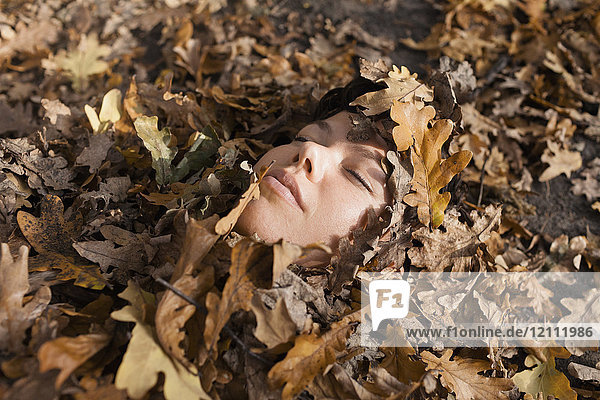 Nahaufnahme einer Frau mit geschlossenen Augen  die mit trockenen Blättern bedeckt ist.