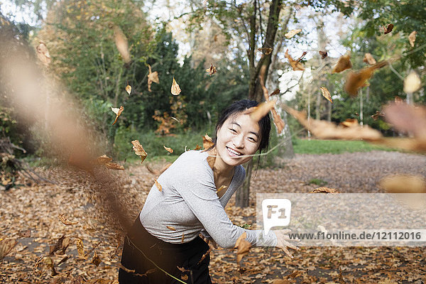 Porträt einer glücklichen Frau  die im Herbst trockene Blätter in die Luft wirft  während sie im Park steht.