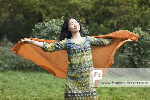 Junge Frau mit orangefarbenem Schal und ausgestreckten Armen im Park