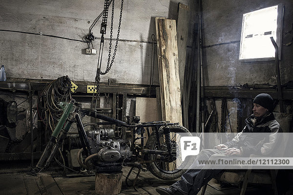 Mechanic sitting in armchair smoking cigarette in workshop with dismantled vintage motorcycle