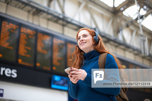 Young woman at train station  wearing headphones  holding smartphone