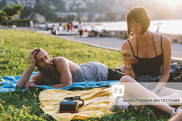 Junges Paar betrachtet Smartphone im Gras am Wasser  Comer See  Lombardei  Italien