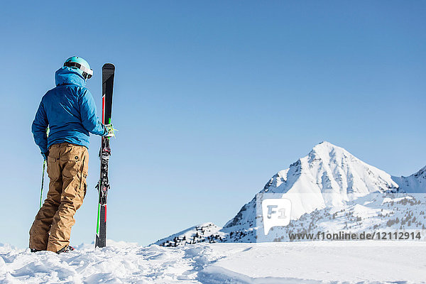Skier standing in snow  holding skis  looking at mountain  rear view