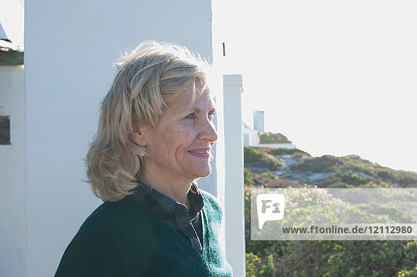 Glückliche blonde  ältere Frau  die von einem sonnenbeschienenen Haus an der Küste nach draußen schaut  Kapstadt  Westkap  Südafrika