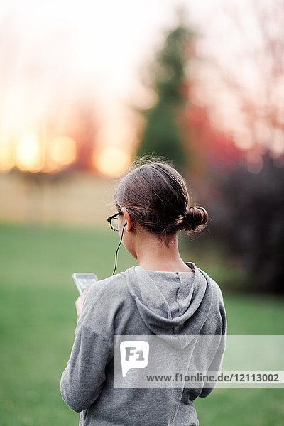 Rückansicht eines Mädchens  das über Kopfhörer Musik hört und auf ein Smartphone im Garten schaut