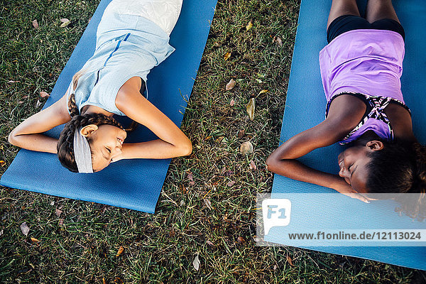 Draufsicht auf zwei Schulmädchen  die auf Yogamatten auf dem Schulsportplatz liegen