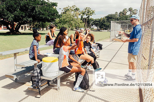 Teacher questioning schoolgirl soccer players on school sports field bench