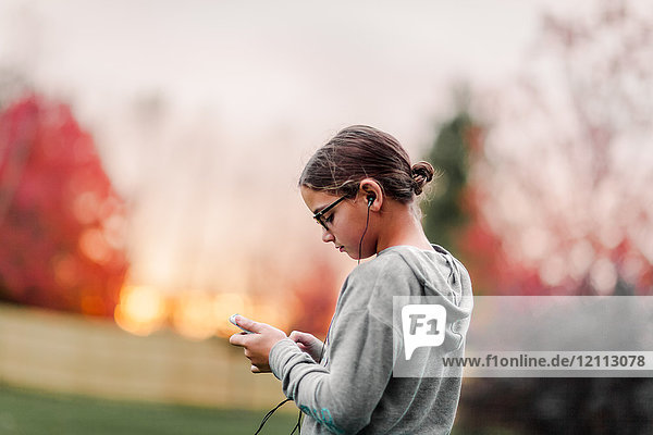 Seitenansicht eines Mädchens  das über Kopfhörer Musik hört und ein Smartphone im Garten betrachtet