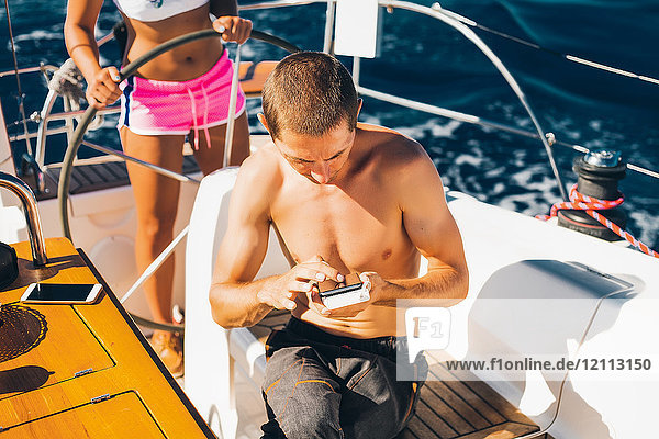 Frau im Bikini steuert Boot  Mann im Vordergrund mit Smartphone