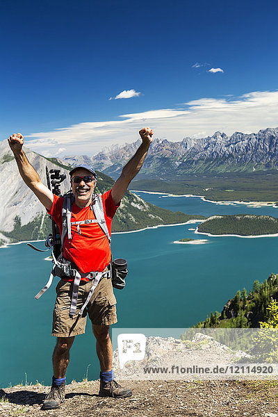Wanderin auf einem Bergkamm mit erhobenen Armen  in der Ferne ein bunter Bergsee und eine Bergkette mit blauem Himmel und Wolken; Kananaskis Country  Alberta  Kanada