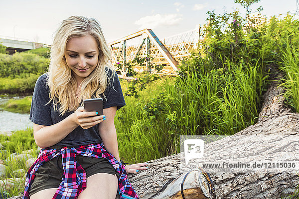 A young woman sits on log in a park using her cell phone with a river and bridge in the background; Edmonton  Alberta  Canada
