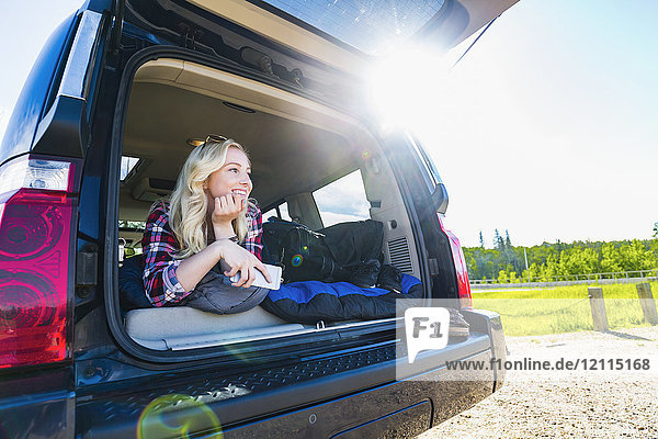 Eine junge Frau auf einer Autoreise liegt auf dem Rücksitz eines Fahrzeugs mit ihrem Handy auf einem Schlafsack und schaut aus der offenen Tür; Edmonton  Alberta  Kanada