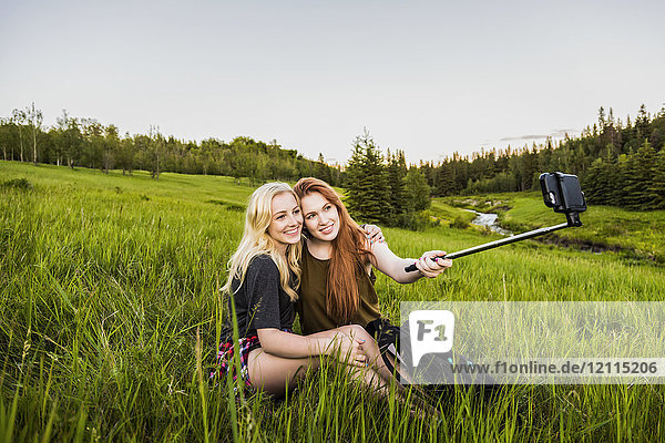 Two girlfriends sitting in a grass field posing for a self-portrait with a selfie stick and smart phone at sunset; Edmonton  Alberta  Canada