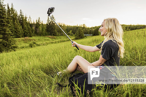 Eine junge Frau mit langen blonden Haaren macht ein Selbstporträt mit einem Selfie-Stick und einem Smartphone  während sie auf einer Wiese in einem Park sitzt; Edmonton  Alberta  Kanada
