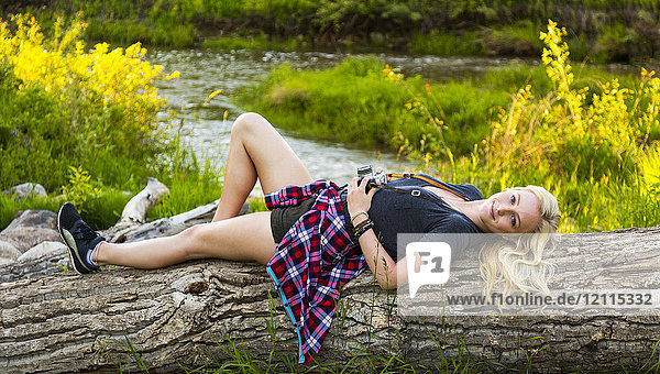 Porträt einer jungen Frau mit langen blonden Haaren  die auf einem Baumstamm in einem Park liegt und eine Kamera hält; Edmonton  Alberta  Kanada