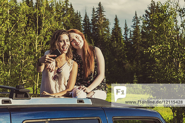 Zwei junge Frauen auf einem Roadtrip stehen auf dem Sonnendach eines Fahrzeugs und machen ein Selbstporträt mit einem Smartphone; Edmonton  Alberta  Kanada