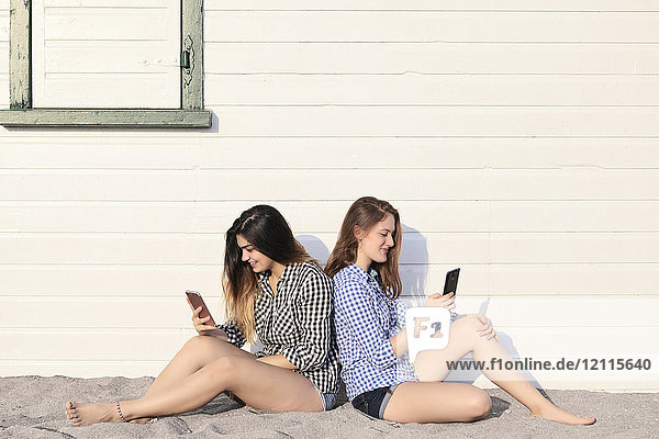 Two teenage girls sitting in the sand against a building looking at their smart phones together  Woodbine Beach; Toronto  Ontario  Canada