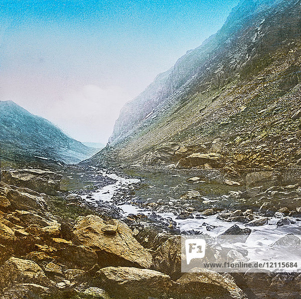 Magic Lantern slide circa 1900 hand coloured. created in 1887. A tour of North Wales. 29 Llanberis Pass  from Pont-y-Cromlech —A little higher up  and we are tempted to declare that this is certainly the wildest and most awe-inspiring spot we have ever been in.“ What lonely magnificence stretches around ! Each sight how sublime ; and how awful each sound ! All hushed and serene  as a region of dreams  The mountains repose ’mid the roar of the streams. On our left is a tremendous piece of rock  known as the Cromlech  which has been thrown down from the side of Glyder Fawr. From a little bridge here we get this view looking down the Pass  and also a better idea of the wildness of the glen  with its almost perpendicular sides. A little further the summit of the Pass is reached  which is appropriately named Gorphwysfa  i.e.  the resting place. From here the finest ascent of Snowdon is made.
