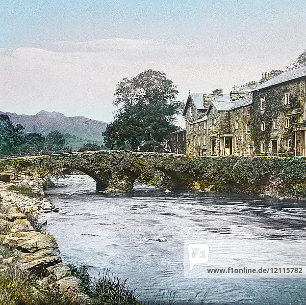 Magic Lantern slide circa 1900 hand coloured. created in 1887. A tour of North Wales. Beddgelert Bridge —Here we get a view of a portion of this very picturesque little village  with its ivy-covered bridge. Beddgelert is situated at the junc¬tion of three vales  and is closely surrounded by mountains. The river Glaslyn  seen in the view  is formed by the union of the streams which flow through Nant Gwynnant and Nant Colwvn respectively. The surrounding scenery is very beautiful and diversified  and the tourist will do well to make a short stay here.
