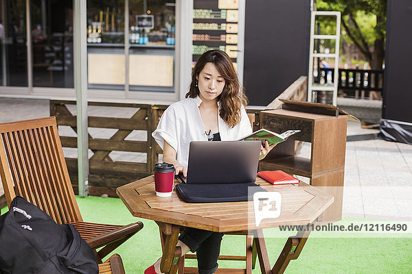 Frau mit schwarzen Haaren und weißem Hemd  die in einem Straßencafé vor einem Laptop am Tisch sitzt und arbeitet.