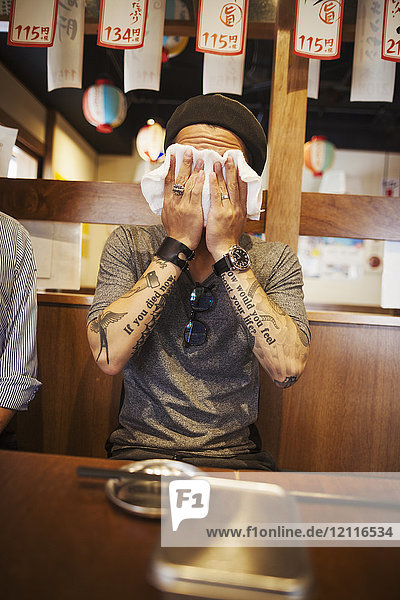 Mann mit tätowierten Armen sitzt an einem Tisch in einem Restaurant und wischt sich das Gesicht mit einem nassen Handtuch ab.