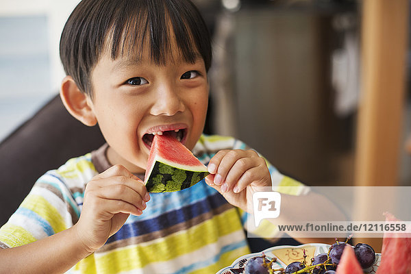 Nahaufnahme eines Jungen mit schwarzen Haaren  der ein gestreiftes T-Shirt trägt und eine Scheibe Wassermelone isst.