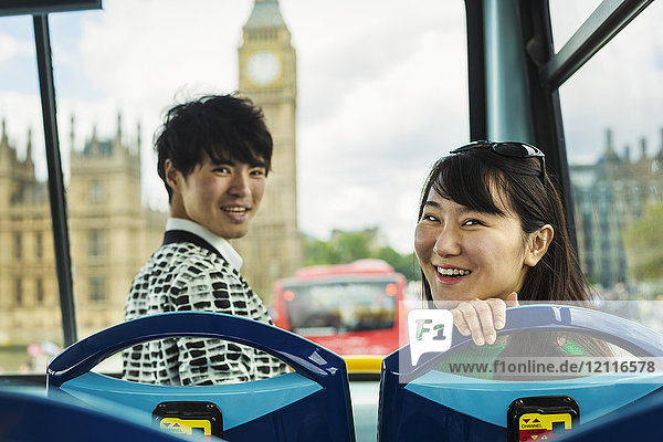 Lächelnder Mann und Frau mit schwarzen Haaren sitzen vorne auf dem Dach eines Doppeldeckerbusses in London und schauen in die Kamera  das Parlamentsgebäude und den Big Ben im Hintergrund.