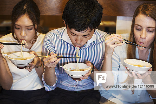 Hochwinkelaufnahme von drei Personen  die nebeneinander an einem Tisch in einem Restaurant sitzen und mit Stäbchen aus Schüsseln essen.