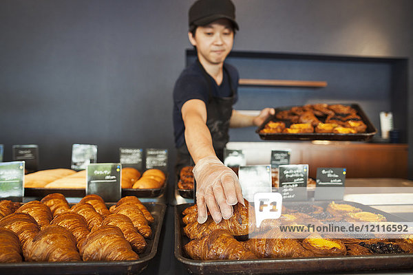 Mann  der in einer Bäckerei arbeitet und frisch gebackene Croissants auf große Bleche legt.