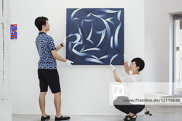Man in blue shirt and woman wearing white shirt hanging modern painting on white wall in art gallery.