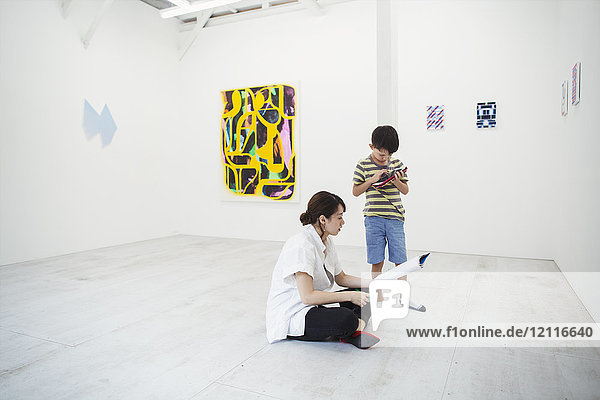 Woman with black hair wearing white shirt sitting on floor in art gallery with pen and paper  boy standing beside her.