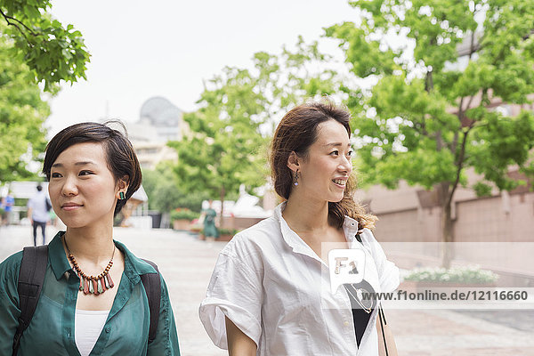 Zwei Frauen mit schwarzen Haaren in weiß-grünem Hemd gehen lächelnd die Straße entlang.
