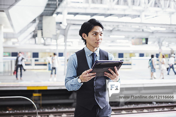 Businessman wearing blue shirt and vest standing on train station platform  holding digital tablet.