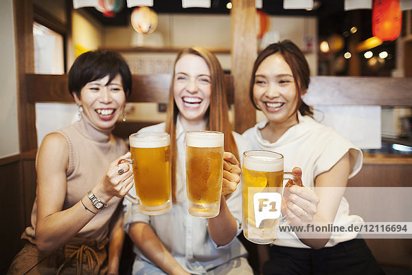 Three women sitting sidy by side at a table in a restaurant  holding large glasses with beer.