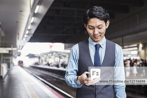 Geschäftsmann in blauem Hemd und Weste steht auf dem Bahnsteig des Bahnhofs und hält ein Mobiltelefon in der Hand.