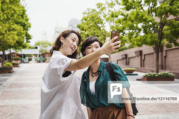 Zwei Frauen mit schwarzen Haaren in weißem und grünem Hemd stehen im Freien  nehmen Selfie mit Handy und lächeln.