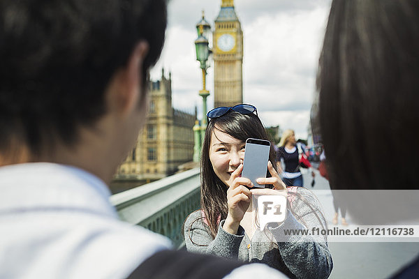 Lächelnde Frau mit schwarzen Haaren  die ein Paar mit einem Smartphone fotografiert  das auf der Westminster Bridge über die Themse in London steht  mit dem Houses of Parliament und Big Ben im Hintergrund.