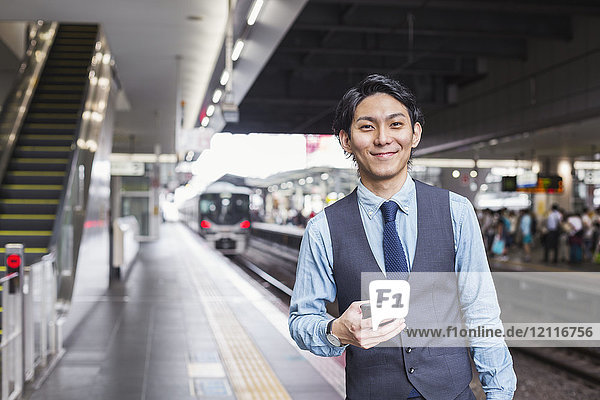 Businessman wearing blue shirt and vest standing on train station platform  holding mobile phone  looking at camera.