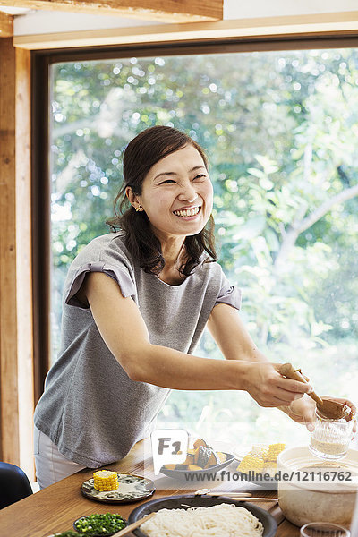 Lächelnde Frau mit schwarzen Haaren steht an einem Tisch mit Schüsseln mit Essen  in der Hand eine Holzkelle.