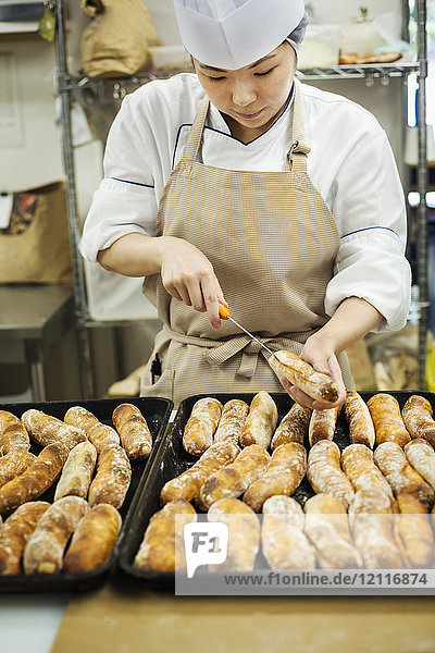 Frau mit Kochmütze und Kochschürze  die in einer Bäckerei arbeitet und frisch gebackene Brötchen auf großen Blechen schneidet.