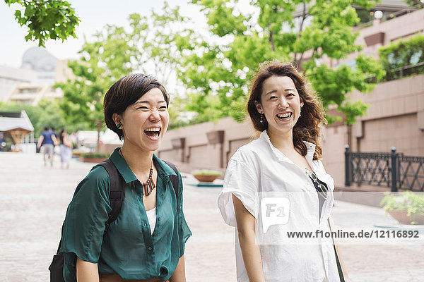 Two women with black hair wearing white and green shirt walking along street  smiling.