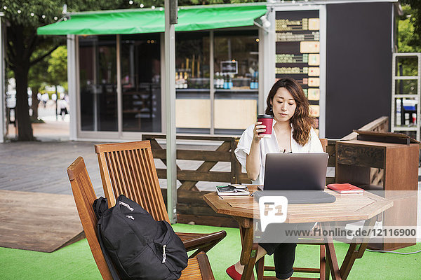Woman with black hair wearing white shirt sitting in front of laptop at table in a street cafe  working.
