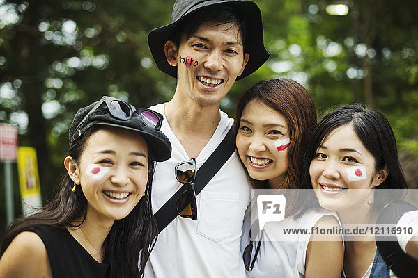 Ein lächelnder Mann und drei junge Frauen stehen im Freien  die Gesichter mit japanischen Flaggen bemalt  in die Kamera blickend.