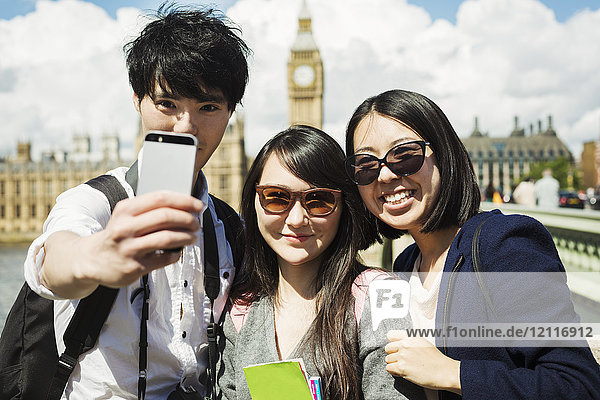 Lächelnder Mann und zwei Frauen mit schwarzen Haaren  die sich mit einem Smartphone selbstständig machen  stehen auf der Westminster Bridge über die Themse  London  mit dem Houses of Parliament und Big Ben im Hintergrund.