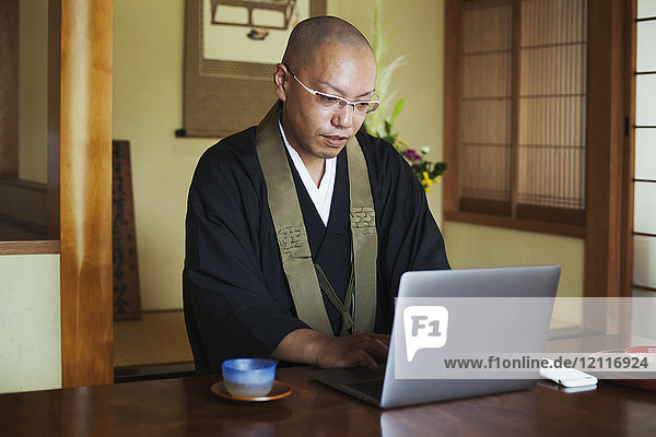 Buddhistischer Mönch mit rasiertem Kopf in schwarzer Robe  der drinnen an einem Tisch sitzt und einen Laptop-Computer benutzt.