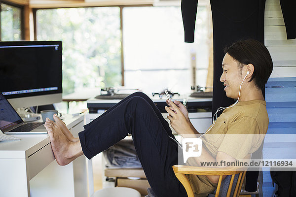 Seitenansicht eines Mannes  der drinnen an einem Schreibtisch mit Computer sitzt  Beine hochgelagert  barfuß  Handy in der Hand  Kopfhörer tragend.