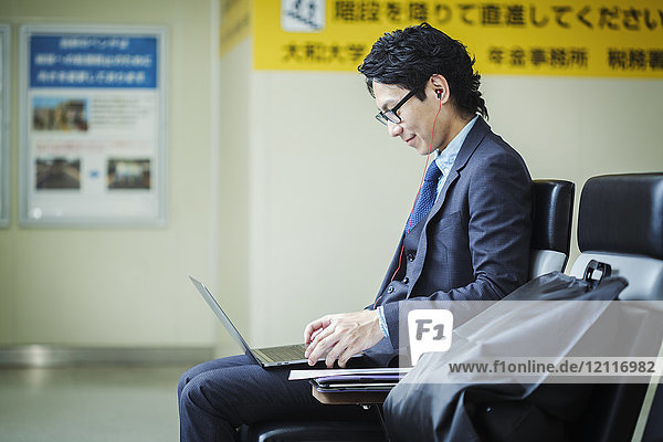 Businessman wearing suit and glasses sitting at train station working on laptop.