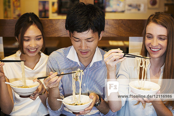 Drei lächelnde Menschen sitzen nebeneinander an einem Tisch in einem Restaurant und essen mit Stäbchen aus Schalen.