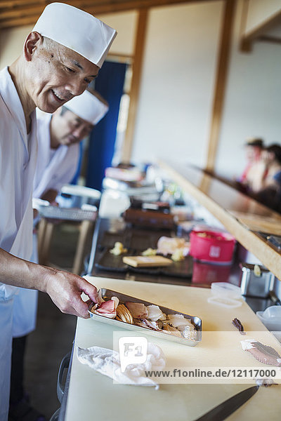 Zwei Köche arbeiten an einer Theke in einem japanischen Sushi-Restaurant und halten ein Metalltablett mit frischem Fisch und Meeresfrüchten in der Hand.