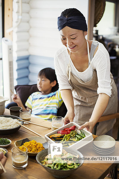 Lächelnde Frau mit Schürze  die Schüsseln mit Salat und Gemüse auf den Tisch stellt  im Hintergrund sitzt ein Junge.